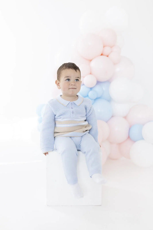 A toddler boy wearing a light blue and white striped long-sleeve polo shirt and matching light blue pants sits on a white block in front of a cluster of pastel pink, white, and light blue balloons.
