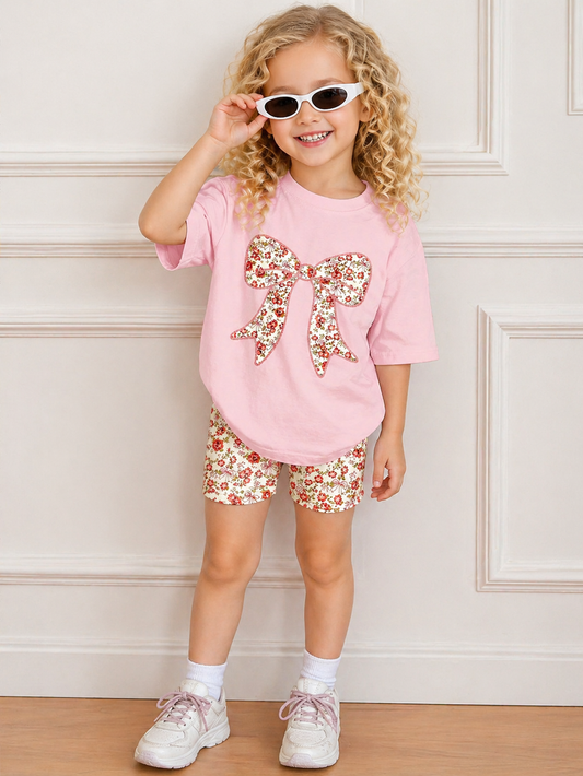 Young girl wearing a pink shirt with a floral bow and matching shorts, standing against a light-colored wall.