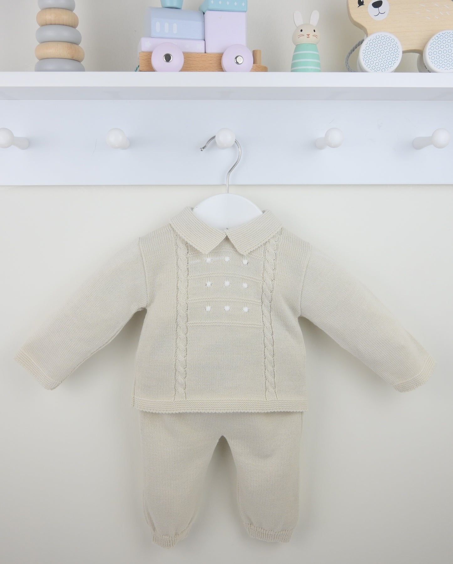Beige baby outfit with shirt and pants on a white surface, toys in the background