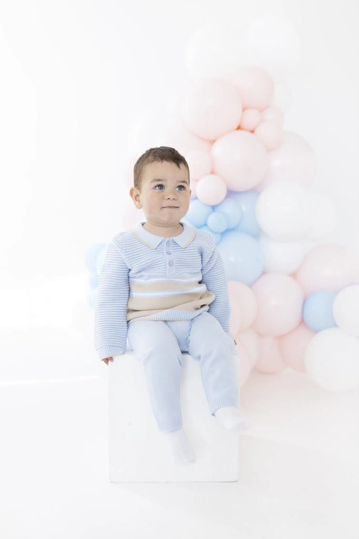 A toddler boy wearing a light blue and white striped long-sleeve polo shirt and matching light blue pants sits on a white block in front of a cluster of pastel pink, white, and light blue balloons.