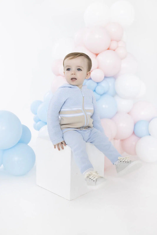 A baby boy wearing a light blue and beige striped zip-up hoodie and matching light blue pants sits on a white block in front of a pastel pink, blue, and white balloon backdrop.