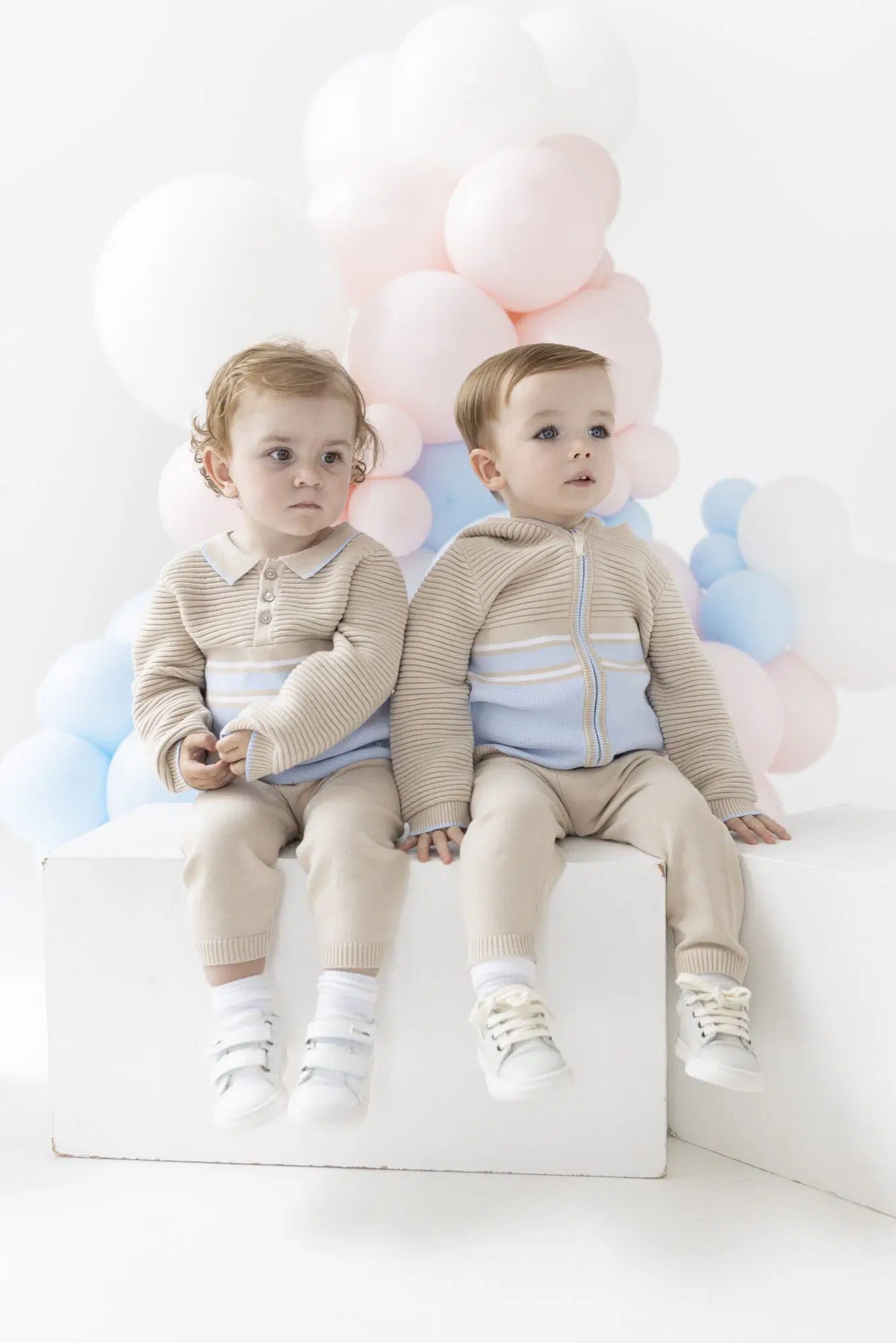 Two toddlers sitting side by side on a white block, wearing matching beige knit outfits with light blue accents. They are posed in front of a soft, cloud-like balloon garland in pastel pink, blue, and white against a clean white studio background.