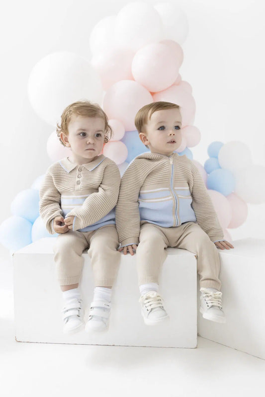 Two toddlers sitting side by side on a white block, wearing matching beige knit outfits with light blue accents. They are posed in front of a soft, cloud-like balloon garland in pastel pink, blue, and white against a clean white studio background.