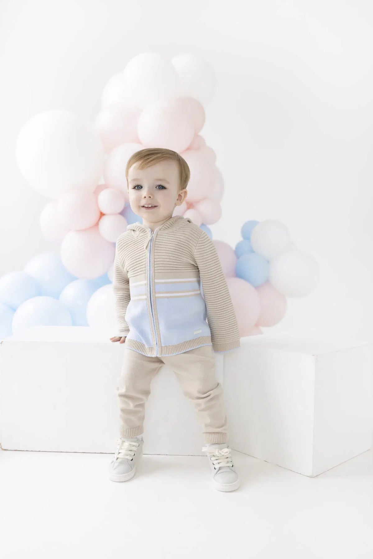 A toddler stands on a white platform wearing a beige and light blue knit zip-up hoodie with matching beige pants and white sneakers, posed in front of a cluster of pastel pink, white, and blue balloons.