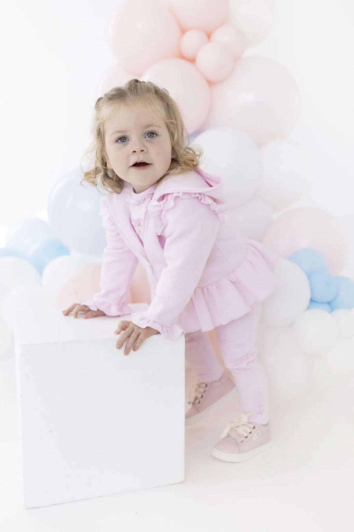 A toddler girl with blonde curly hair wearing a light pink ruffled outfit and pink sneakers, posing on a white block in front of a background of pastel pink, white, and light blue balloons.