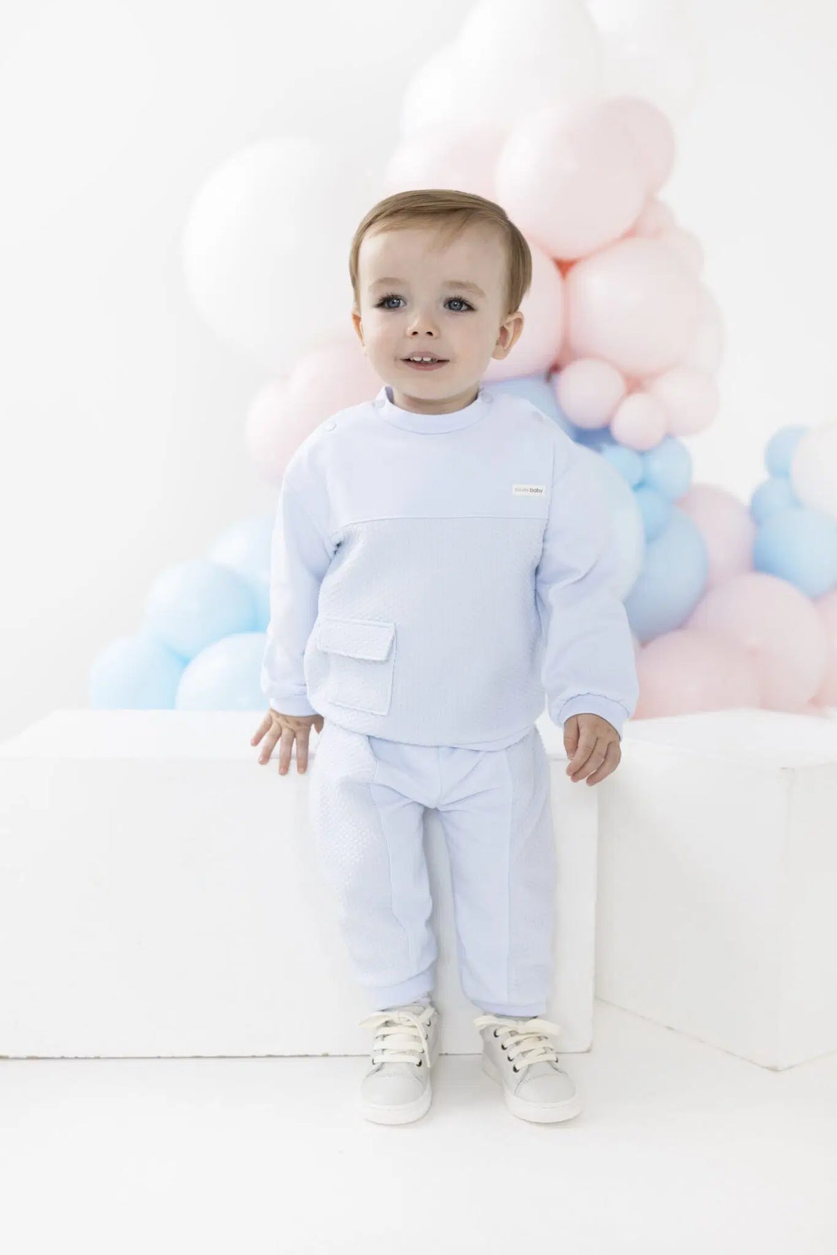 A baby boy wearing a light blue long-sleeve top and matching pants, standing in front of a white studio background decorated with pastel pink, blue, and white balloons.