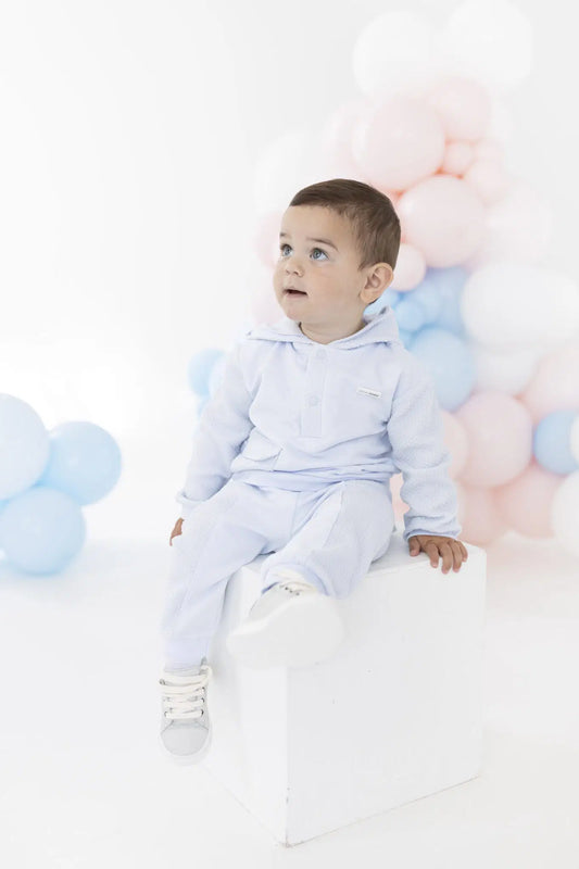 A toddler boy wearing a light blue hooded sweatshirt and matching sweatpants sits on a white block against a white studio background decorated with pastel pink, blue, and white balloons.