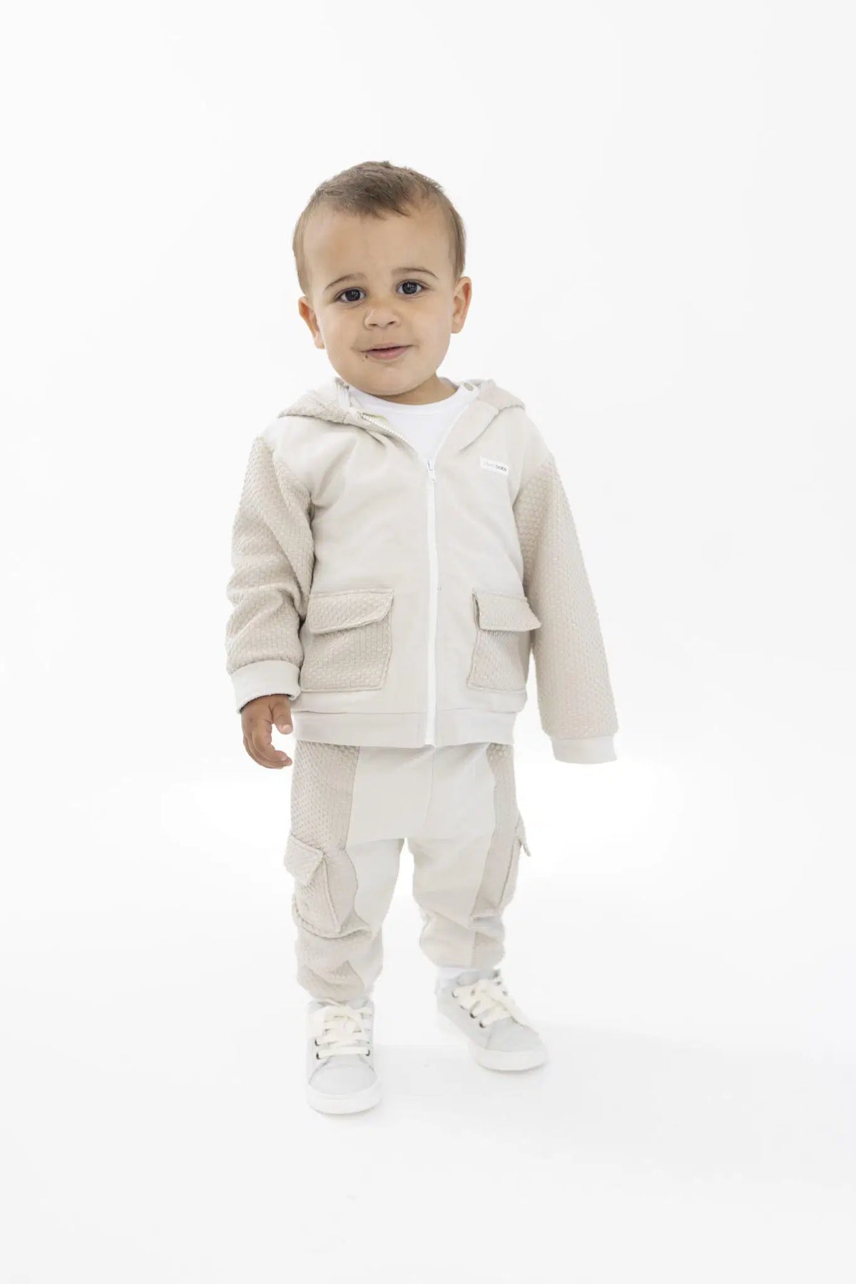 A toddler standing against a plain white studio background, wearing a light beige hooded zip jacket, matching beige pants, and white sneakers.