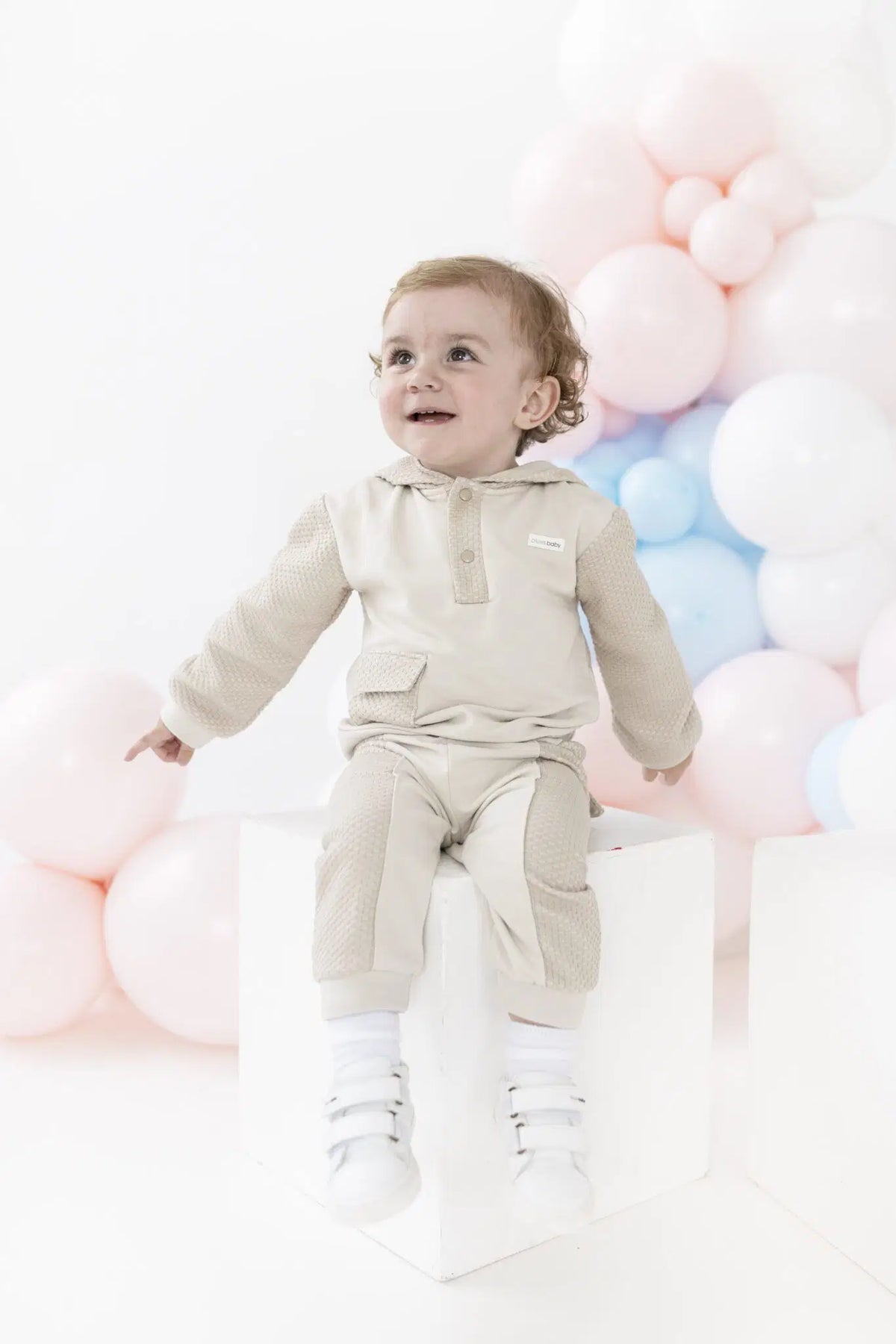 A baby wearing a light beige hooded knit outfit sits on a white block against a white background decorated with pastel pink, blue, and white balloons.