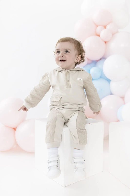 A baby wearing a light beige hooded knit outfit sits on a white block against a white background decorated with pastel pink, blue, and white balloons.