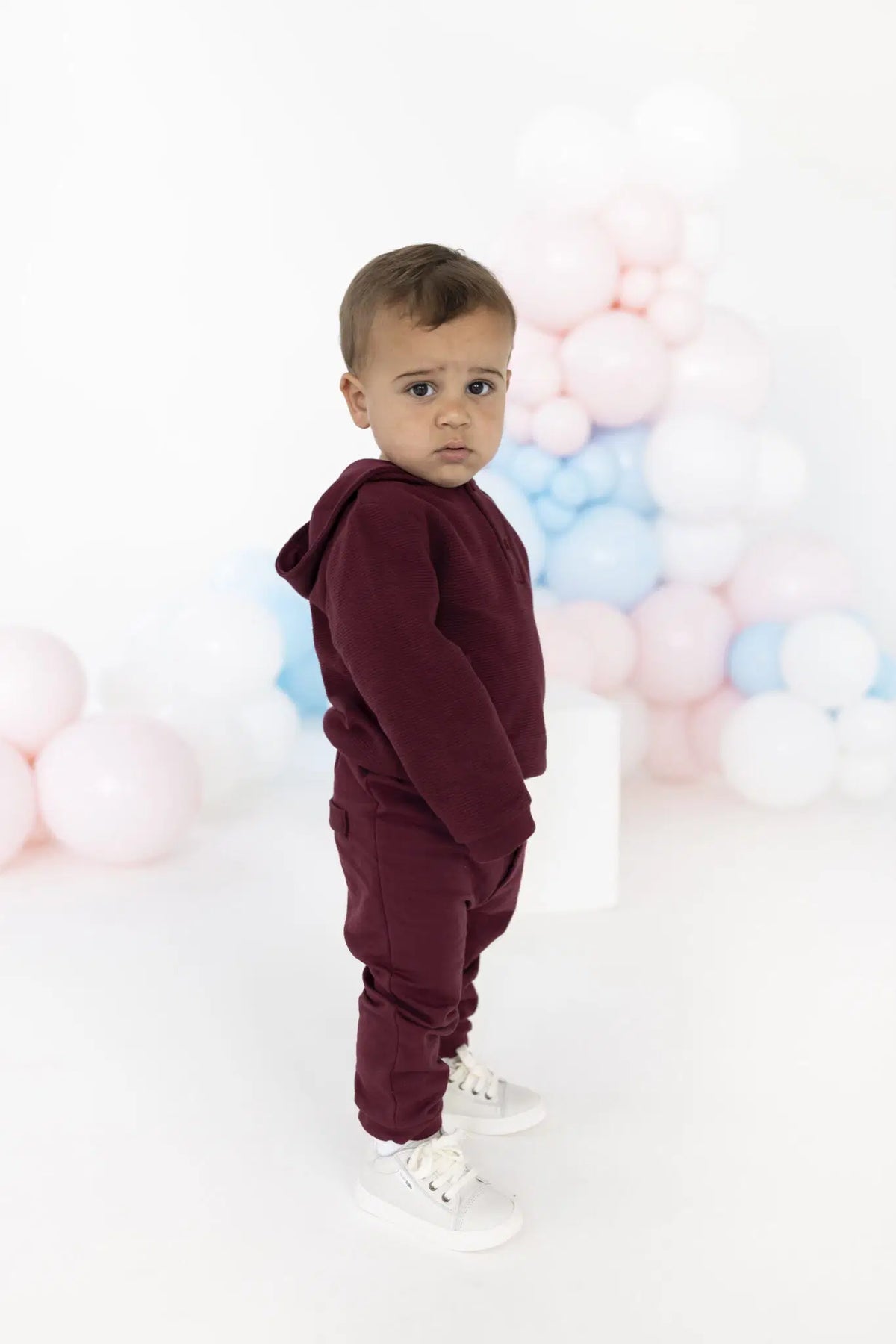 A toddler stands in profile wearing a matching burgundy hooded sweatshirt and jogger pants set, paired with white sneakers, against a white studio background decorated with pastel pink, blue, and white balloons.
