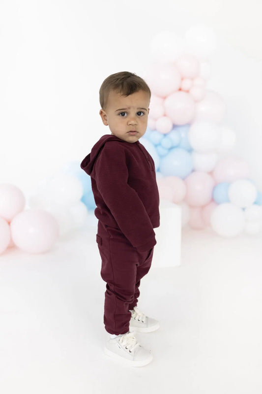 A toddler stands in profile wearing a matching burgundy hooded sweatshirt and jogger pants set, paired with white sneakers, against a white studio background decorated with pastel pink, blue, and white balloons.