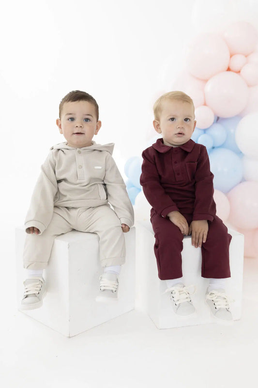 Two toddlers sitting on white pedestals, wearing matching two-piece outfits in beige and burgundy. The background is a clean white studio setting decorated with pastel pink and blue balloons.