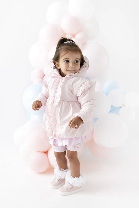 A toddler girl wearing a light pink puffer jacket, matching pink ruffled shorts, and pink fuzzy booties stands in front of a soft pastel balloon backdrop of pink, white, and light blue.