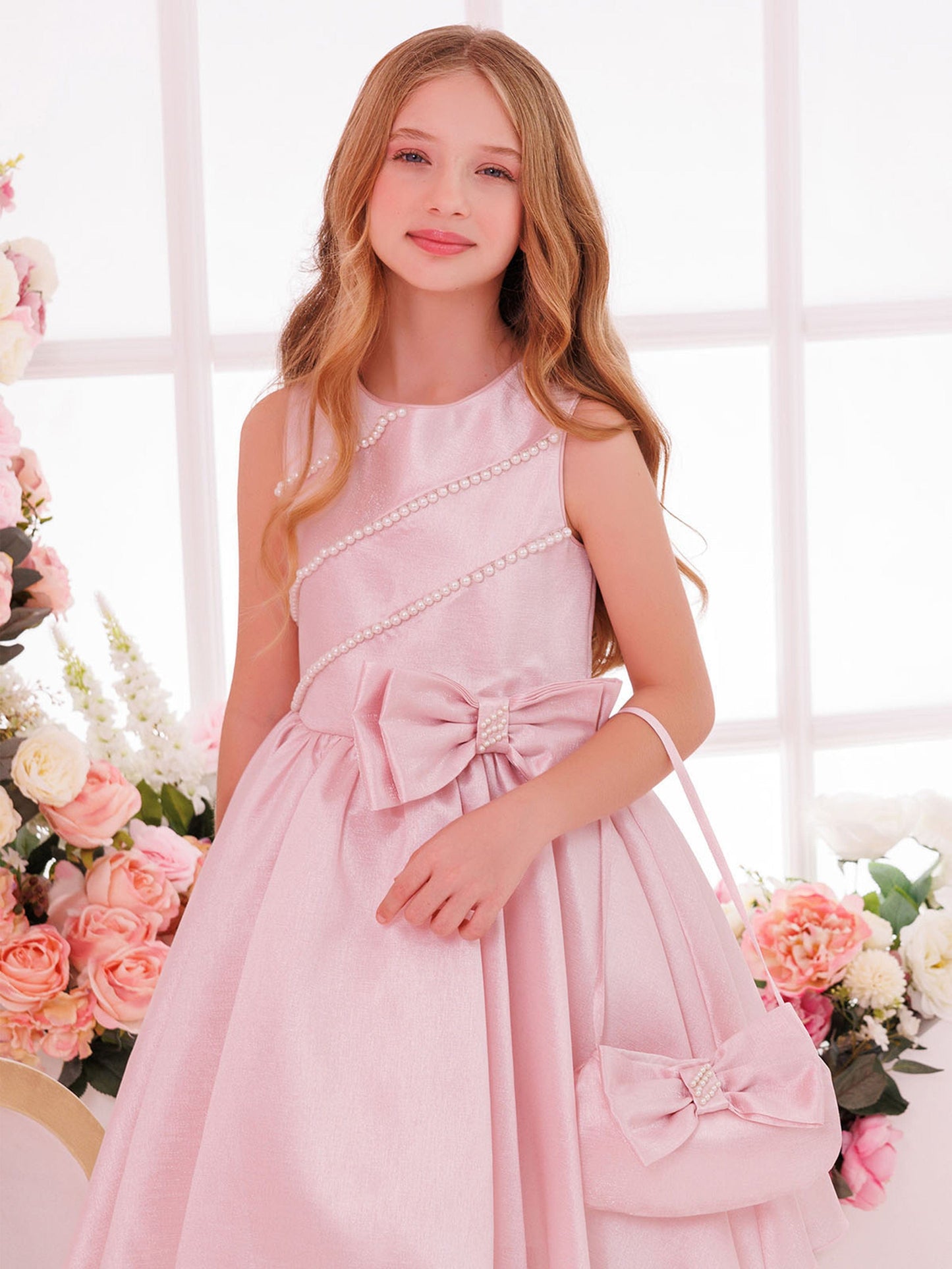 Girls pink satin dress with pearl trim and bow waist, holding mini bag
