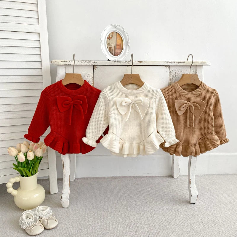 Baby knit pullovers hanging on hooks in red, white, and camel with bow accents