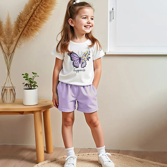 Young girl wearing a white t-shirt with a butterfly design and purple shorts, standing in a room with a wooden table and plant.