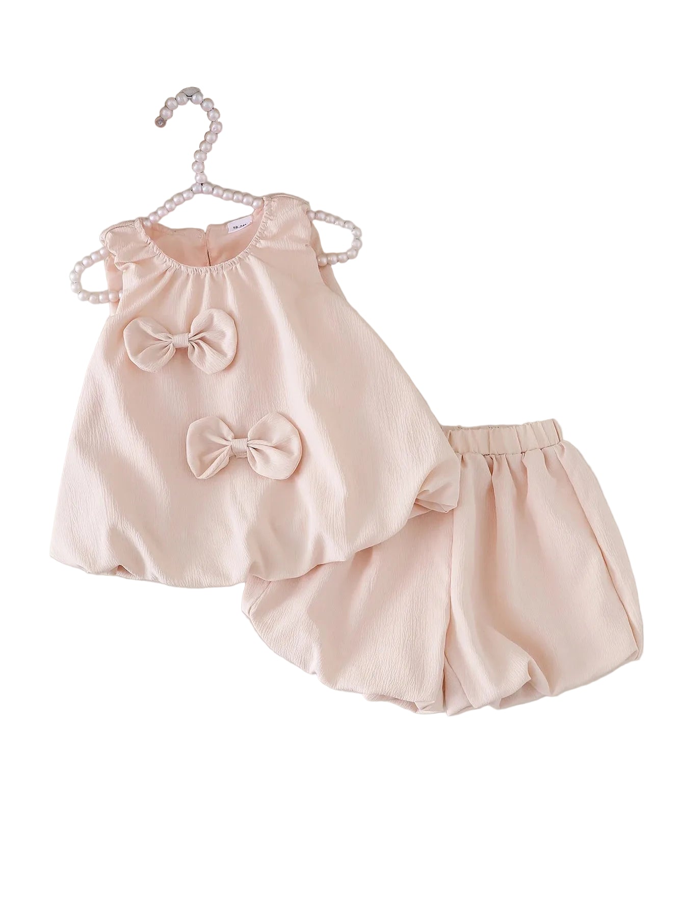 Pink baby outfit with bows on a white surface, accompanied by sandals and flowers.