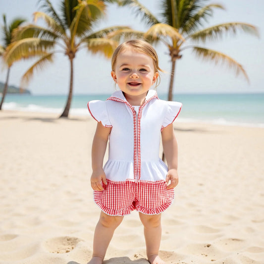 Child in a red and white checkered outfit standing on a sandy beach with palm trees in the background.