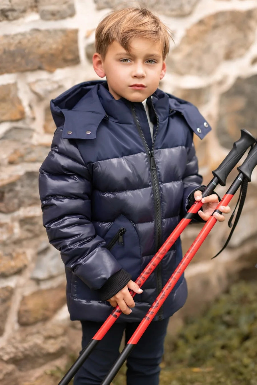 Child wearing a navy puffer jacket holding red ski poles against a stone wall.