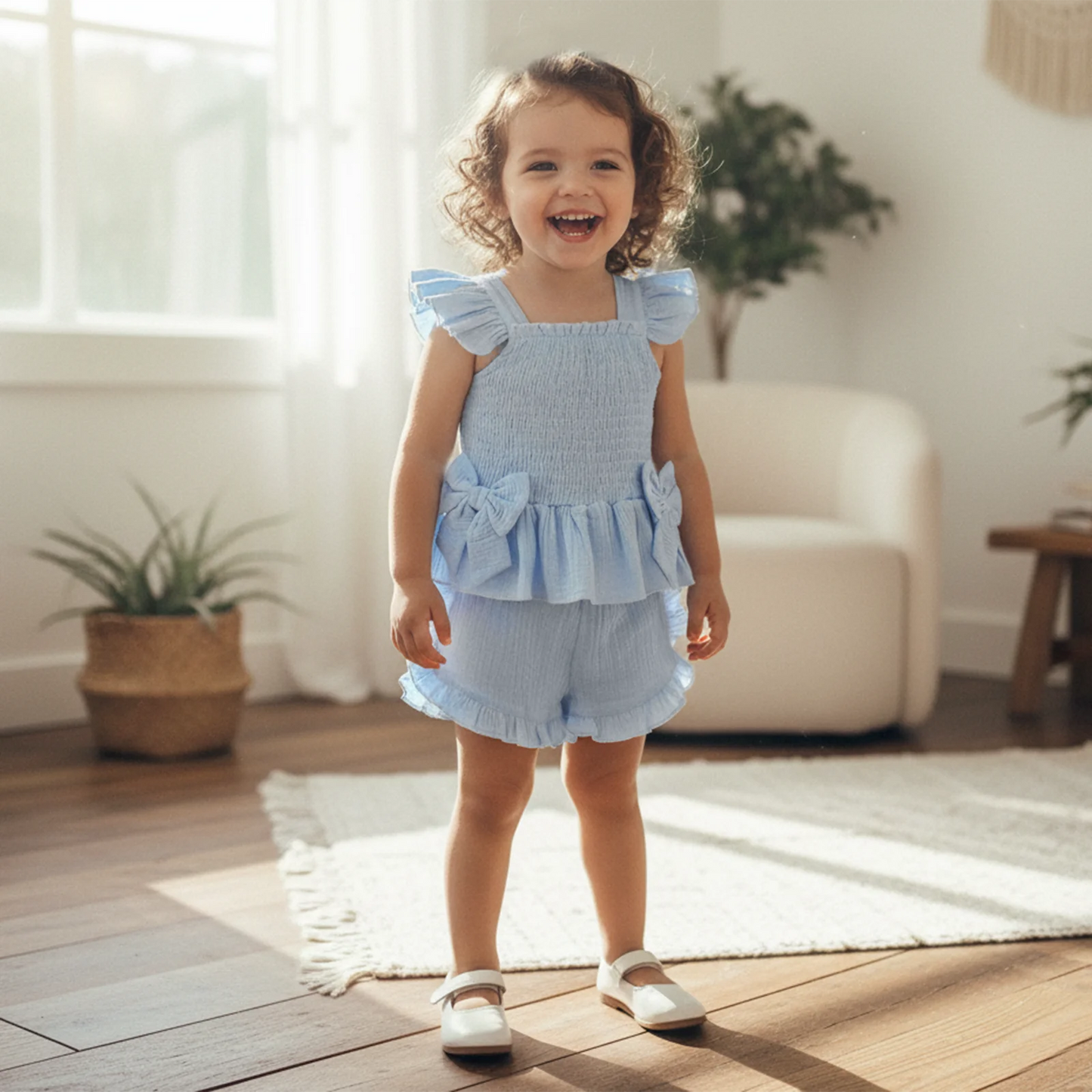 Child wearing a light blue outfit standing in a bright room with plants and furniture.