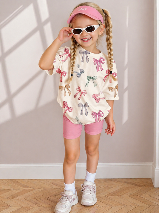 Young girl in a white shirt with pink bows, pink shorts, and sunglasses standing against a light-colored wall.